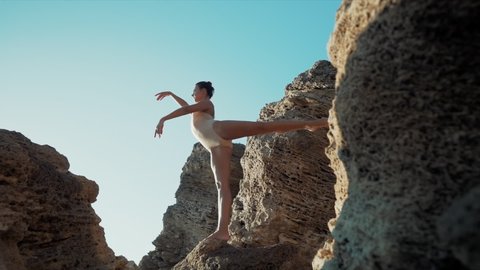 Dancing ballerina among rocks near ocean or sea at sunrise or sunset. Portrait of flexible woman practicing in classic exercises. Concept of tenderness, lightness, art and talent in nature