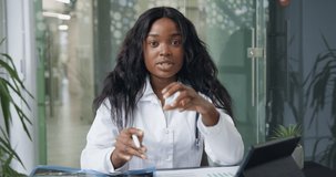 Front view of beautiful confident experienced black-skinned female doctor in white gown with stethoscope which holding video conference and consulting partners or patients, looking into camera