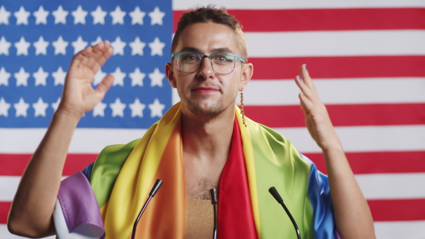 Young gay man with makeup, colored nails and LGBT rainbow flag over his shoulders standing at rostrum against American flag and speaking into microphones during press conference with journalists