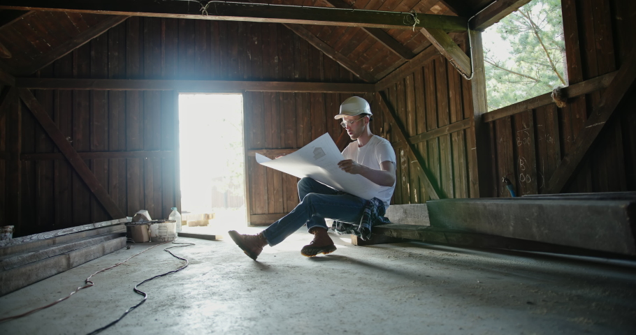 Man in casual clothes and hardhat sitting on boards and checking draft inside timber barn. Male builder examining blueprint of shed