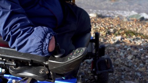 An unidentified caucasian man with amputated two stump hands and legs is controlling his electric wheelchair along the beach.