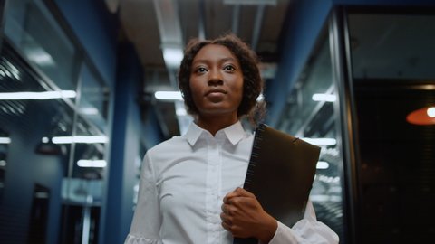 Portrait of focused businesswoman walking on meeting in office corridor. Closeup african american woman walking in corridor. Serious afro business woman going with folder in business center hallway.