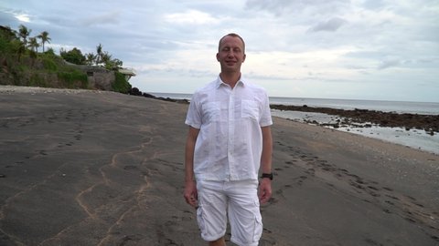 Young man walks on background of sea. A man in a white shirt and shorts walks along the black sand beach. Romantic morning dawn. Media