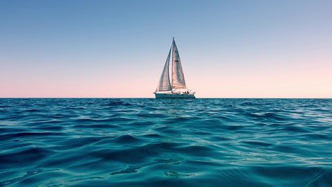 Low-angle sea-level view of small yacht boat sailing in calm open sea at sunset