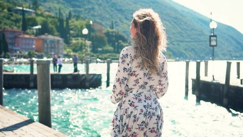 Young woman long blond hair fly in wind white short fluttering dress walk. old town waterfront Riva del Garda colorful houses. Enjoy fabulous view lake Lago di Garda. Trentino Alto Adige region Italy 
