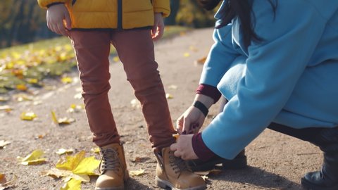 Close up of loving mother helping little boy to put on shoes in park. Babysitter taking care of child and tying shoes outdoors in autumn park.