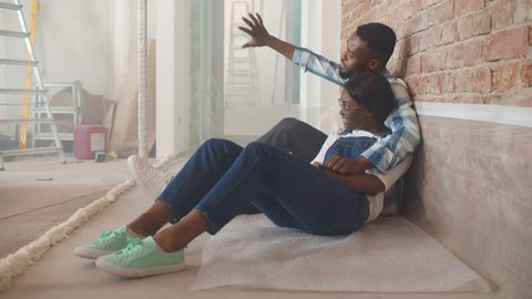 Cheerful black couple talking planning renovation and interior design sitting on floor after moving in new house. Happy afro man and woman discussing redecoration of apartment