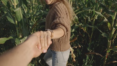 Back or rear view of blonde-haired girl in brown hat leading young man hand through tall green thickets of corn. Happy young woman making her man follow her. Slow motion and Follow me concept.