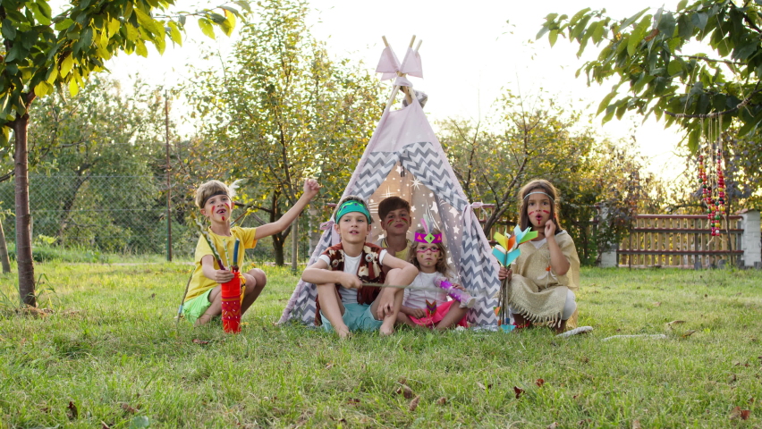 Five Children Play Indians. Three Boys And Two Girls Are Sitting In Tent Outdoors With Painted Faces And Arrows In Their Hands. Some Have Feathers On Their Heads. One Boy Is Holding Stick. He iIs