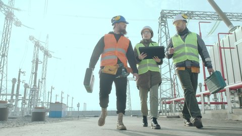 A team of energy engineers, two men and a woman in overalls, helmets, carrying a tool case, walk through the substation against the backdrop of a powerful high-voltage transformer and high-voltage