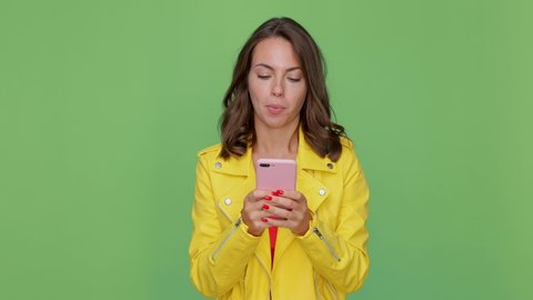 Confused puzzled young woman in yellow leather jacket isolated on green background studio. People lifestyle concept. Using mobile phone typing sms message ask what spreading hands showing thumb down