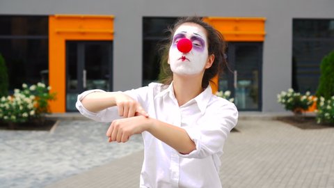Funny young beautiful clown shows on watch and stare to camera.