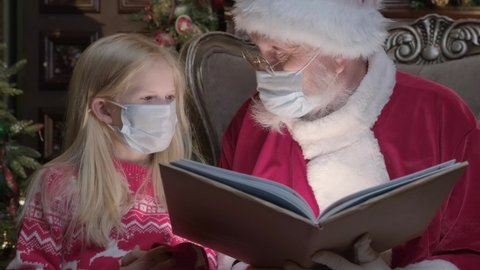 Authentic Santa Claus in protective mask from virus reads magic book with blonde girl. Light from book shines on face of Girl and Santa reading Christmas stories in dark room