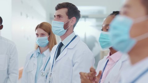 Multi-race professional medical doctors and assistants wearing protective masks against coronavirus applauding on team conference business employment meeting in corridor. Cooperation and success.