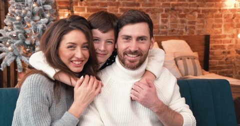 Cute Family Portrait, little Boy is hugging his Daddy and Mom. Christmas Mood, Christmas Decorations on the Backgroud, Family wearing Christmas Sweaters. Celebration. Cozy Evening at Home.