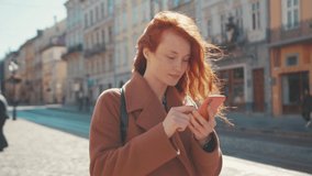 Amused enthusiastic young ginger woman with open mouth holding smartphone in hands. Surprised female blogger collects likes after posting video in social media, city street in background. Popularity