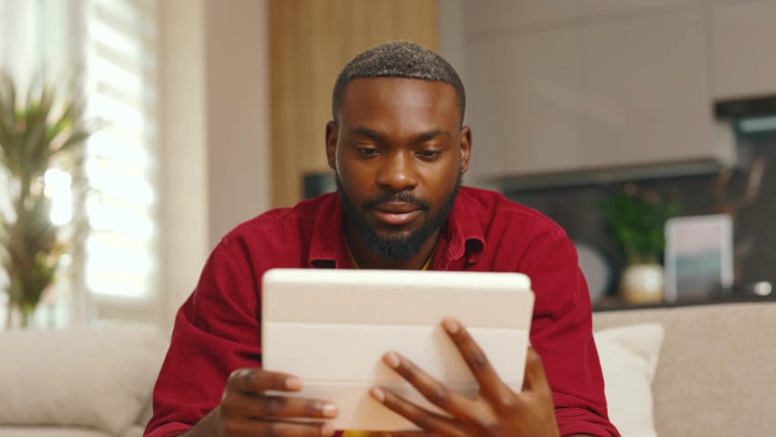 Happy african american young man using digital tablet computer at home sitting on sofa. Handsome smiling guy. Technology thinking black apartment online indoor. Slow motion | Shutterstock HD Video #1061966389