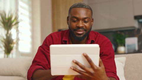 Happy african american young man using digital tablet computer at home sitting on sofa. Handsome smiling guy. Technology thinking black apartment online indoor. Slow motion