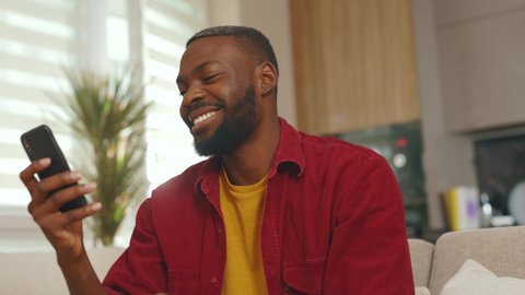 Cute happy young african american man use phone smiling read good news sit on sofa in home. Communication in social media online relax work app. Handsome guy. Internet. Slow motion