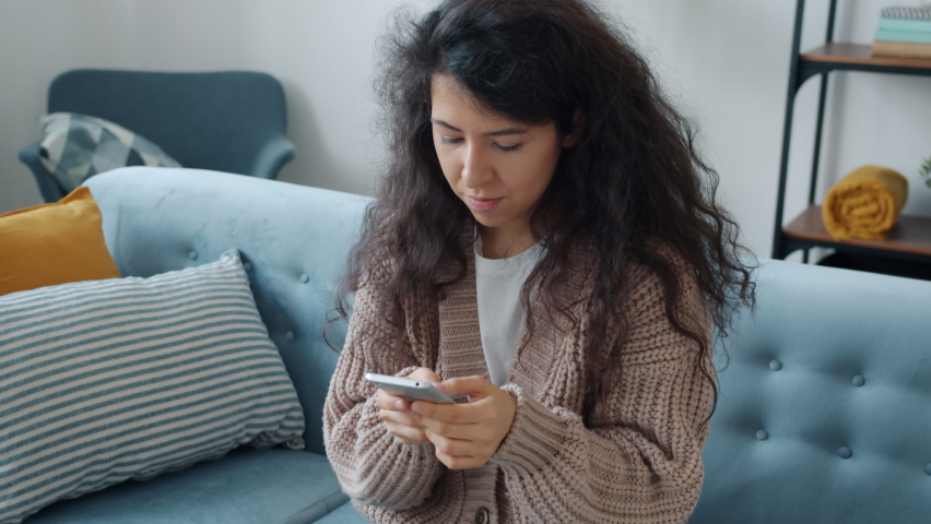 Hapy mixed race girl is using smartphone touching screen then smiling getting good news showing thumbs-up hand gesture sitting on sofa at home