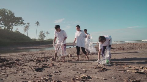 Arc shot of four indonesian ecology activists with trash bags walking along sandy coastline collecting garbage, waste and plastic for further reuse helping environment