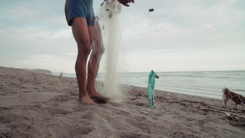 Low-section footage of local indonesian man at coastline getting out plastic trash out of net with dog running nearby