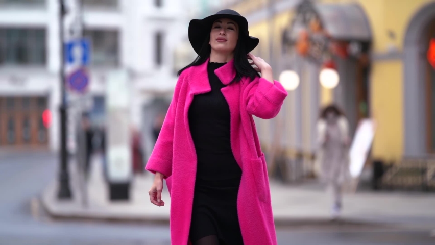 A stylish black-haired woman in a pink coat and black hat walks and poses on a blurry city street background. | Shutterstock HD Video #1061973688