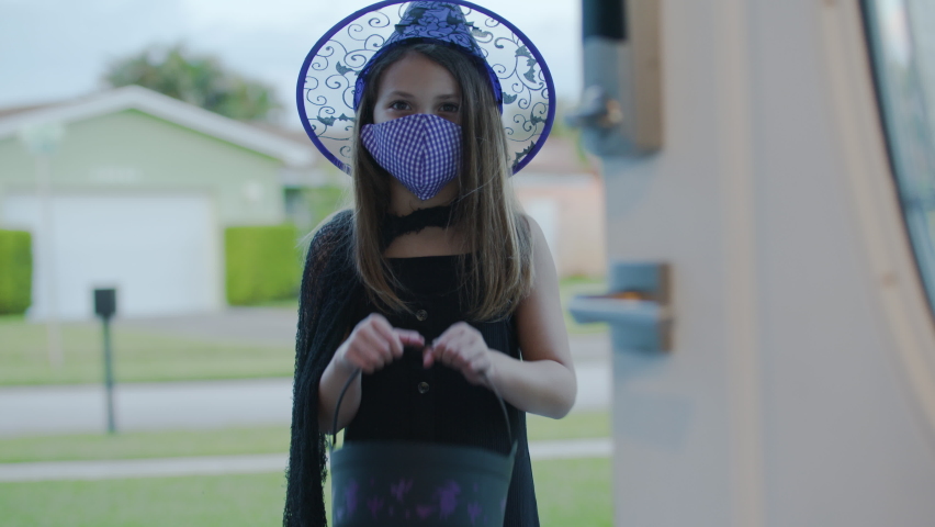Little girl celebrates a Covid-19 Safe Halloween by trick-or-treating while wearing a mask. Door opens to reveal girl in witch costume wearing a mask for protection. | Shutterstock HD Video #1061977267