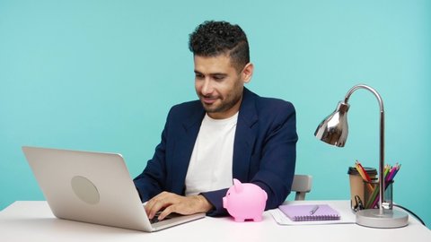 Smiling positive businessman showing piggy bank and thumbs up sitting on workplace in office, satisfied with investments to future. Indoor studio shot isolated on blue background