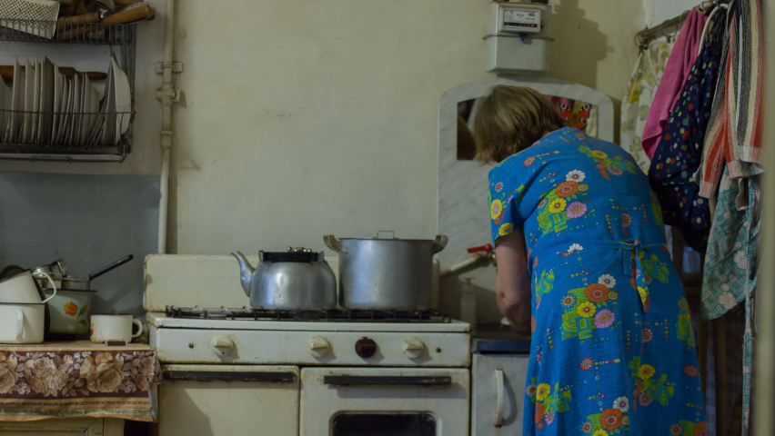 Elderly woman washes the dishes and lights the fire in the old gas stove and puts a kettle to boil, in the kitchen in an old rural house