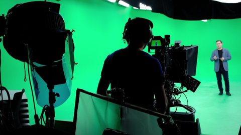 The cameraman follows the announcer in a professional green screen studio. The professional looks all the shooting equipment; lights, camera, green screen, monitors, filters