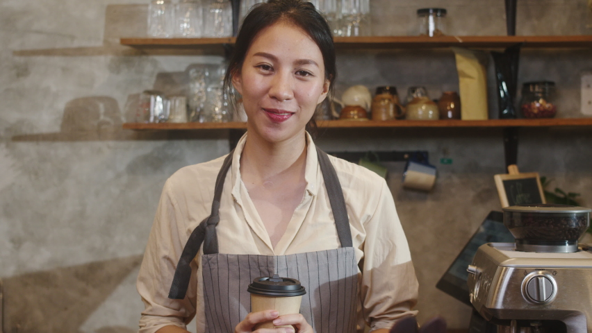 Portrait young Asian lady barista waitress holding coffee cup feeling happy at urban cafe. Asia small business owner girl in apron relax toothy smile looking to camera stand at counter in coffee shop. | Shutterstock HD Video #1061988610
