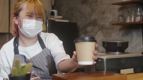 Young Asia female barista wear face mask serving take away hot coffee paper cup to consumer at cafe. Owner small business, lifestyle new normal after corona virus and social distancing concept.