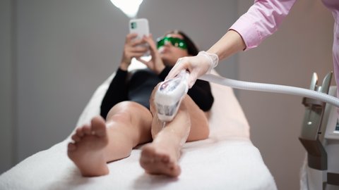 Attractive beautician doing laser hair removal on legs of beautiful young woman who is lying on medical couch and texting on social networks by phone in a beauty salon. Cosmitology and spa concept