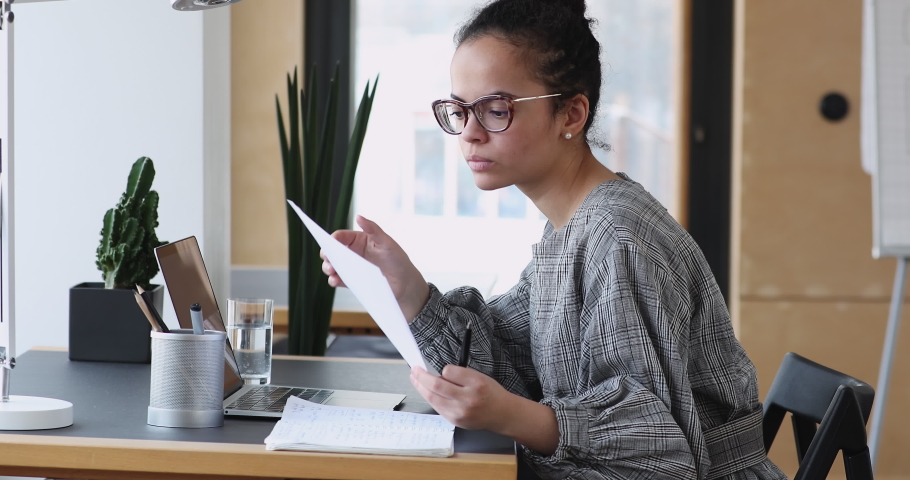 Focused young african ethnicity woman sitting at workplace, reading information analyzing paper documents. Concentrated skilled mixed race employee secretary checking report or contract in office. Royalty-Free Stock Footage #1061988922