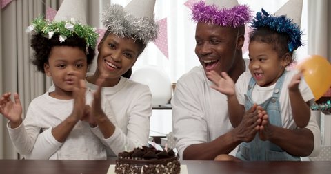 Happy black parents and little cute children african american family in party hat celebrating birthday on online video call during lockdown, birthday boy blowing candles on cake making wish.