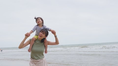 Asian family on vacation. Beautiful mother carrying little baby girl on shoulder walking on the beach together with happy and smile. Soft touch, love care, protection of mom and child relationship.