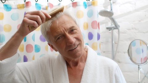 Senior man in bathrobe combing hair in bathroom in morning. Portrait of elderly male brushing hair with comb after showing doing morning routing preparing for work