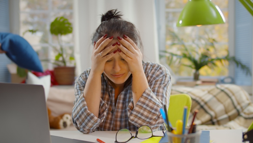 Tired mother with stress working on laptop at home with noisy children on background. Young exhausted woman putting off glasses and holding head sitting at desk with kids playing on background | Shutterstock HD Video #1062002062