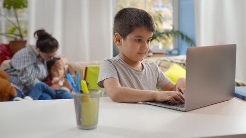 Happy student boy with laptop computer learning at home with mother and little brother relaxing on couch on background. Preteen kid playing video game on computer at home