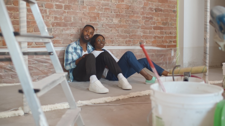 African couple sitting on floor in empty room of new home planning design. Portrait of cute afro-american couple relaxing on floor in redecorating apartment Royalty-Free Stock Footage #1062002182