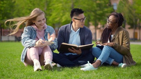 Multiethnic university students sitting and reading book on campus grass. Portrait of three diverse classmates relaxing in park studying together reading textbook and writing notes