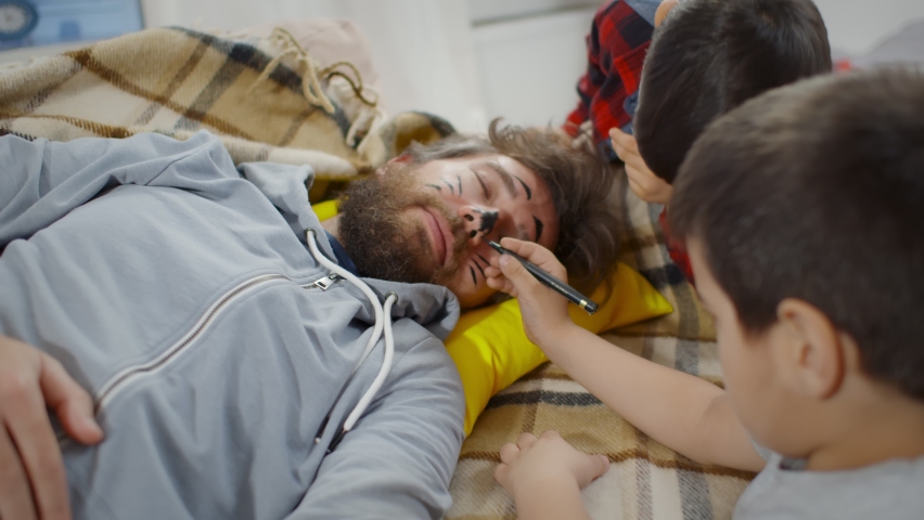 Mischievous boys painting face of father sleeping on couch at home. Adorable preschool kids drawing with marker on sleeping dad face and having fun. Childhood concept
