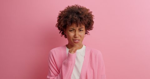 Young dissatisfied ethnic curly haired female model waves hand near nose feels unpleasant smell squints because of stench doesnt like something disgusting isolated over pink studio background.