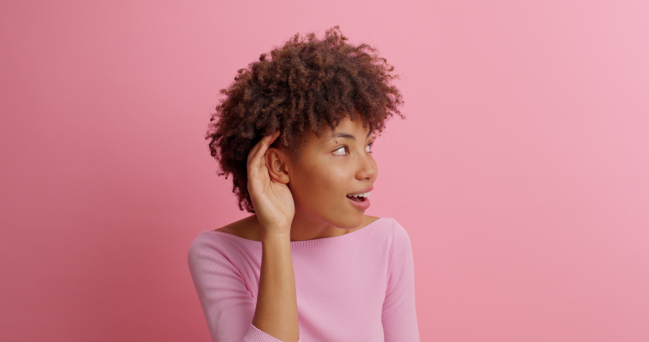 Cute curious woman keeps hand near ear tries to overhear something very interesting listens somebodys secret looks away likes collecting rumors poses against pink background. Eavesdropping concept | Shutterstock HD Video #1062002629