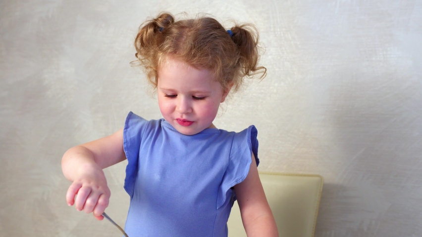 A little Caucasian girl eats something tasty with a spoon and licks her lips with pleasure.