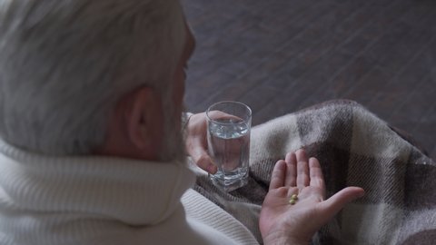 Elderly man taking pills, drinking water in living room at home.