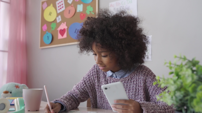 African american child kid school girl holding phone distance learning class using mobile application, watching online tutoring lesson, video calling in app making notes studying at home in classroom. | Shutterstock HD Video #1062010603