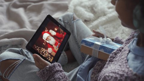 Happy african american kid girl talking to virtual santa claus saying New Year greetings to child on video conference online call chat using tablet sitting on bed opening Chrtistmas present.