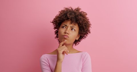 Bewildered confused dark skinned woman has doubts and hesitation about something looks at different sides cant make right decision poses against pink background. Confused Afro American girl.